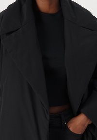 Black oversized jacket with a wide collar, made from smooth fabric. A fitted black top is underneath, paired with high-waisted black pants.