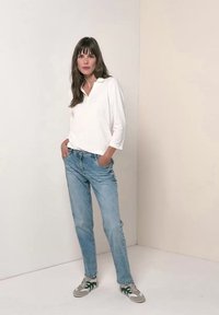 Woman standing with hands in pockets wearing white long-sleeve shirt, light blue jeans, and gray sneakers against plain wall background.
