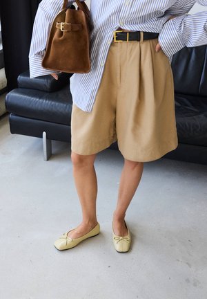 Person wearing beige shorts, striped long-sleeve shirt, holding brown suede bag, and cream ballet flats standing on a light floor.