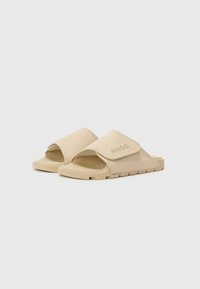 Beige slide sandaler med polstret rem, gummisåle og "HUGO"-logo. Har en bred åpning for enkel av- og påtaking og teksturert fotseng.