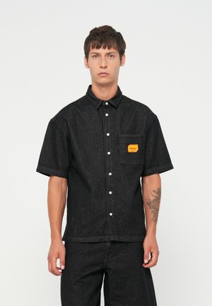 Short-sleeve black denim shirt featuring a button-down front, chest pocket with yellow label, and a boxy fit. Smooth texture and structured design.