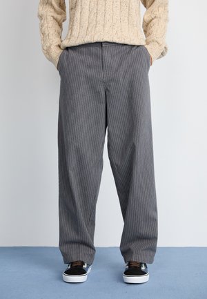 Persona vestida con un suéter de punto beige, pantalones de pierna ancha a rayas grises y zapatillas negras y blancas, de pie sobre un suelo azul.