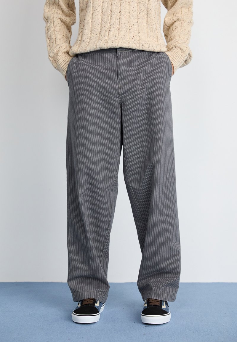 Persona vestida con un suéter de punto beige, pantalones de pierna ancha a rayas grises y zapatillas negras y blancas, de pie sobre un suelo azul.