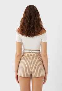 Top bianco off-the-shoulder con bordi scallop abbinato a pantaloni corti beige a vita alta con righe bianche verticali e una cintura.