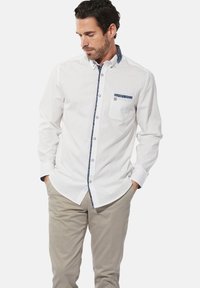 Weißes Hemd mit knöpfbarem Kragen, das einen gemusterten Kragen und eine Tasche aufweist. Glattes Gewebe und taillierter Schnitt, mit kontrastierenden blauen Akzenten entlang der Knöpfe.