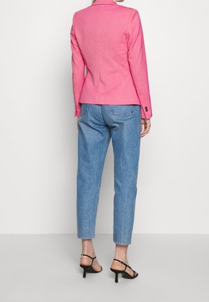 Kvinde iført en tætsiddende lyserød blazer, blå højtaljede jeans og sorte sandaler med hæl, stående med ryggen til mod en hvid baggrund.