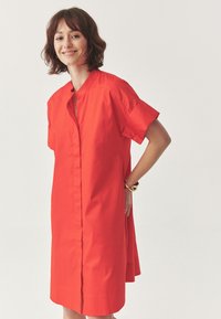 Red, short-sleeve shirt dress with a buttoned front, collar, and side slits; made from a smooth fabric with a solid color.