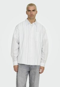 Witte overhemden met blauwe en grijze verticale strepen, met een borstzak en een relaxte pasvorm. Draagt met lichtgrijze jeans.