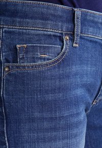 Jeans in denim blu scuro con un design classico, caratterizzati da una tasca frontale con cucitura visibile e rivetto metallico, texture liscia e una leggera scoloritura.