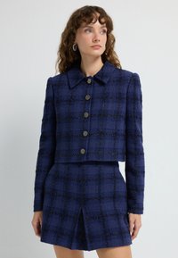 Navyblå tweedjacka med en kortad design, stor krage och sex runda knappar, i kombination med en matchande kjol med en slits fram.