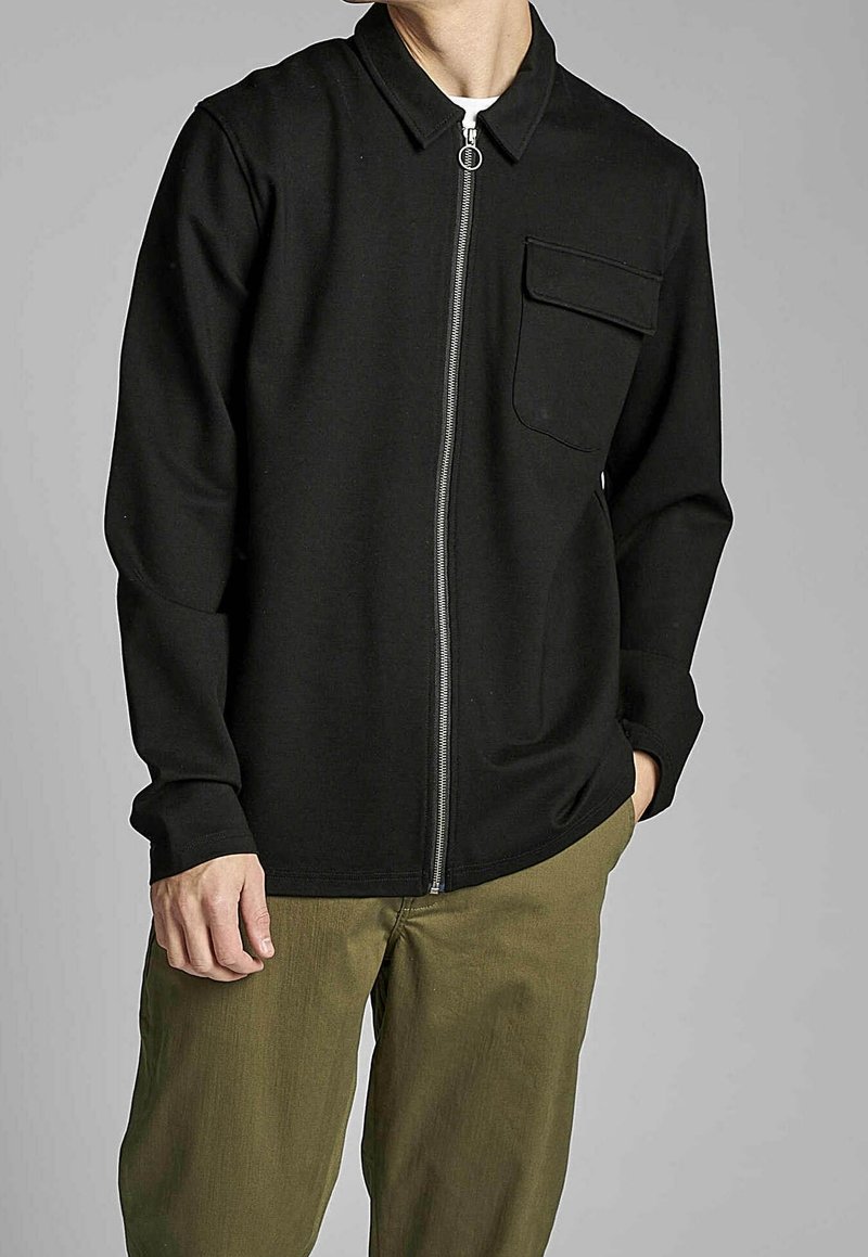 Chemise noire à fermeture éclair avec col, poche avant et manches longues, associée à un pantalon vert olive, dotée d'une texture lisse.