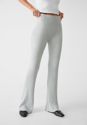 Persoon draagt een lichtgrijze, geribbelde, hooggetailleerde broek met uitlopende pijpen en zwarte instappers, staand tegen een effen witte achtergrond.