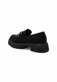 Mocasines de ante negro con suela de goma gruesa, que presentan una hebilla decorativa en el delantero y un diseño de puntera redondeada. Superficie superior texturizada.