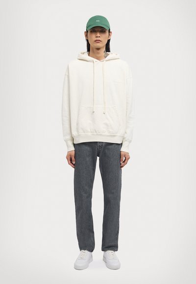 Cream hoodie with front pocket, gray jeans, and white sneakers. Model wears a green cap. Hood features drawstrings; relaxed fit design.