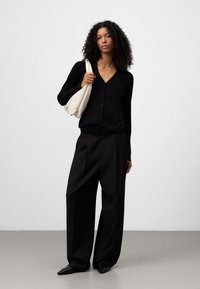 Zwarte, lange mouwen cardigan met een V-hals en knopen, gecombineerd met zwarte wijde broek en een crèmekleurige schoudertas.