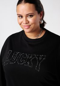 Sweatshirt noir avec un design scintillant "LUCKY" sur le devant, fabriqué en matériau doux avec un col ras du cou et des poignets côtelés.