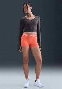 Melna ilgiem piedurknēm crop top, oranžas Nike Pro šortus ar baltu logotipa joslu. Modelis stāv baltās sporta kurpēs uz pelēka fona.
