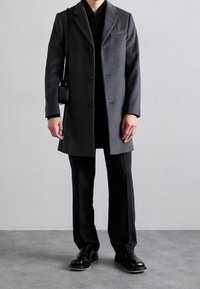 Cappotto grigio in lana con design strutturato, monopetto e rever a sciala. Indossato sopra una maglia nera e abbinato a pantaloni e scarpe neri.