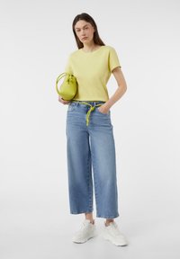 Žlutý strukturovaný pletený top a světlemodré široké džíny, s uzlovým zeleným opaskem a malou zelenou kabelkou. Bílé tenisky dokončují celý outfit.