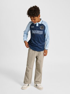 Junge mit lockigem Haar trägt ein marineblaues und hellblaues langärmeliges Sporttrikot, beige Hose und weiße Sneakers, steht und schaut nach unten.