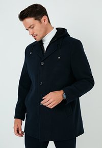 Chaqueta con capucha de color azul oscuro, hecha de tejido suave, con cierre de botones, dos bolsillos frontales y un corte relajado con un dobladillo ligeramente curvado.