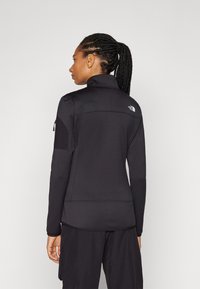 Black athletic zip-up jacket with a high collar, long sleeves, and textured fabric. Features a small logo on the upper back and side pocket.