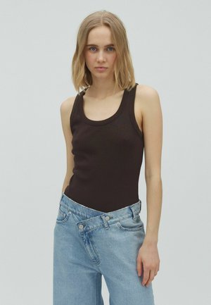 Jeune femme aux cheveux blonds mi-longs porte un haut noir sans manches et un jean large bleu clair avec la taille déboutonnée.