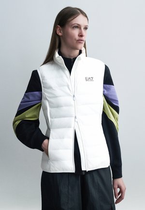 Junge Frau mit langen Haaren, die eine weiße gesteppte Weste über einem schwarzen Sweatshirt mit lilafarbenen und grünen Ärmeln trägt, steht mit einer Hand in der Tasche.