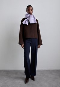 Chaqueta corta marrón con mangas anchas, combinada con pantalones de pierna ancha azul marino y una bufanda de canalé lila clara. Tacones marrones de punta.