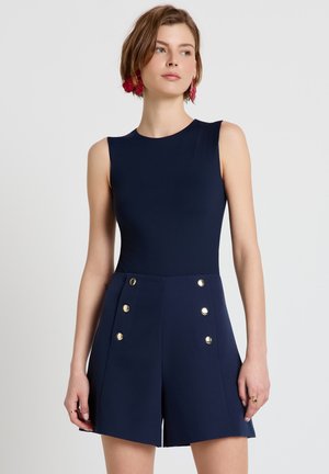 Frau trägt ärmelloses marineblaues Oberteil und passende hoch taillierte Shorts mit goldenen Knöpfen, steht vor einem schlichten hellgrauen Hintergrund.