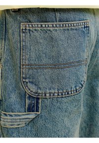 Nærbilde av blå denimjeans som viser en rektangulær, sydd baklomme med forsterkede sømmer og en beltehempe under.