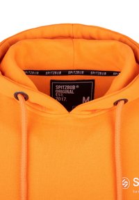 Oranje hoodie met capuchon, voorzien van een trekkoord, etiket met "SPITZBUB" label en witte logovoorziening aan de rechteronderkant. Glad stoffen textuur.