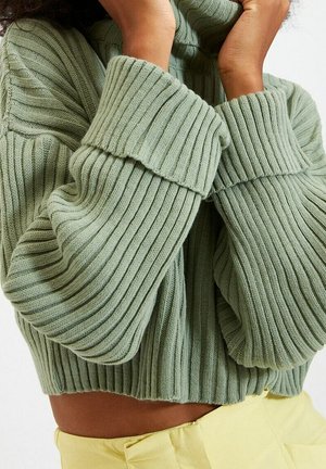 Osoba ubrana w jasnobeżowy prążkowany sweter z długimi rękawami, w połączeniu z wysokim stanem jasnżółtymi spodniami, ręce przy szyi.