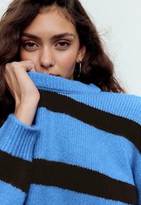 Plavi i crni prugasti pleteni džemper s visokim ovratnikom, rebrastim manžetama i mekom teksturom, djelomično prekrivajući lice s raskošnom valovitom kosom.