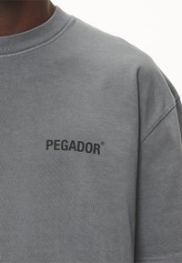 Primer plano de una camiseta de cuello redondo gris con el texto negro "PEGADOR" impreso en el pecho, llevada por una persona de piel oscura.