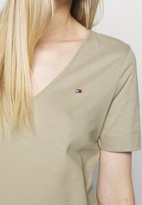 Camiseta de cuello en V de color beige, hecha de algodón suave, que presenta un pequeño logo bordado en rojo, blanco y azul en el lado izquierdo del pecho. Manga corta.