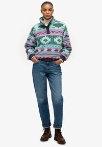 Fleece pullover in blauwgroen en lavendel met geometrische patronen, gecombineerd met blauwe spijkerbroek en bruine veterlaarzen, voorzien van een hoge kraag en knopen.