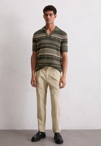 Groene gebreide polo met horizontale strepen; korte mouwen, V-hals en textuurruggen. Gecombineerd met beige broek en zwarte schoenen.