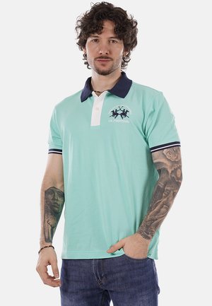 Uomo con capelli ricci che indossa una polo azzurro chiaro con colletto scuro, tatuaggi su entrambe le braccia e jeans blu, in piedi davanti a uno sfondo bianco.