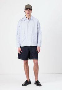 Lys blå stripete skjorte med knapper foran, kombinert med mørke denimshorts. Svarte lærsko og en mønstret caps fullfører antrekket.