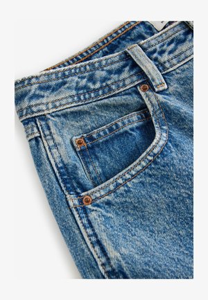 Mėlynos džinsinės kelnės su nežymiu dėvėjimu, vario detalėmis ir oranžine siūle. Yra priekinė kišenė ir tekstūruotas audinio paviršius.