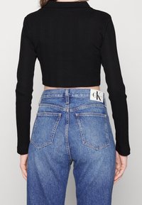 Kvinna i en svart långärmad cropped top och högmidjade blå Calvin Klein-jeans, vänd bort från kameran.
