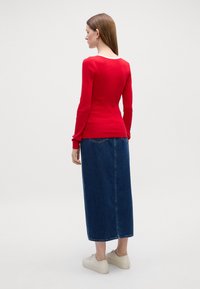 Rød langermet topp med ribbet struktur og rund hals, kombinert med et denim maxi-skjørt. Modellen har på seg beige sneakers.