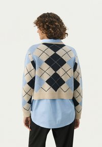 Skrócony sweter w kolorze niebieskim, kremowym i granatowym z wzorem argyle. Dzianinowa tekstura i ściągacz na dole. Nosi się go na jasnoniebieskiej koszuli.