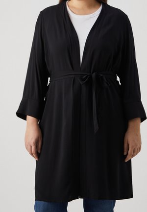 Nainen, joka pukeutunut mustaan pitkähihainen avoimella edustalla olevaan neuletakkiin, jossa on vyötärösidonta, yllä valkoinen paita ja siniset farkut.