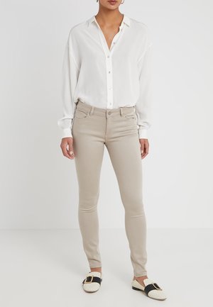 Woman wearing a loose white button-up shirt, beige slim-fit pants, and white loafers with black straps and gold buckles, standing against a plain background.