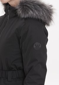 Svart vadderad jacka med en avtagbar grå pälskrage, elastisk midja och en rund präglad logotyp på vänster ärm.