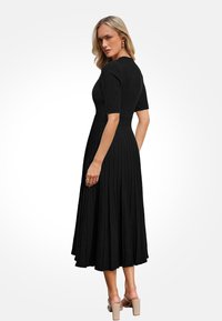 Zwarte gebreide jurk met korte mouwen, strakke bodice en een uitlopende geplooide rok. Het model draagt doorzichtige sandaaltjes met hakken.