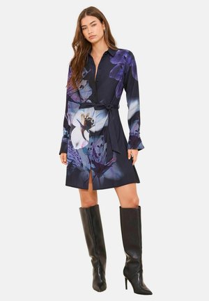 Hemdblusenkleid aus dunklem Stoff mit großem lila Blumenmuster, durchgehender Knopfleiste und Gürteldetail in der Taille. Mit langen Ärmeln und einem Oberschenkelschlitz gearbeitet. Die Stiefel sind kniehoch und schwarz.