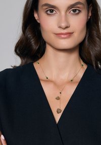 Złoty naszyjnik z zielonymi kamieniami i dwoma akcentami w kształcie liści, zawieszony na delikatnym łańcuszku, o kształcie litery Y.